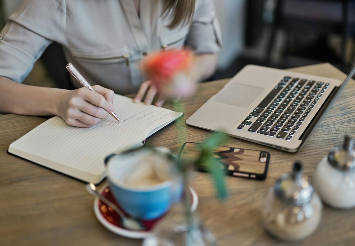 rédactrice écrivant à la main dans un carnet à côté d'un ordinateur portable et d'une tasse de café